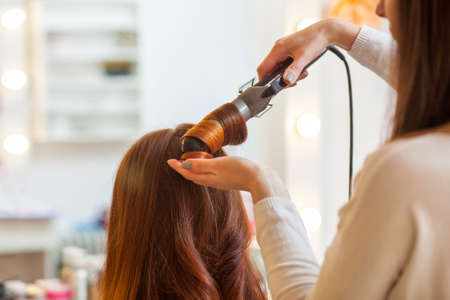 Hairdresser makes hairstyle girl with long red hair in a beauty salon. Create curls with curling irons. Professional hair care.の写真素材