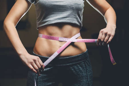 Slim girl bodybuilder centimeter measuring her waist in a gym. Sports concept, fat burning and a healthy lifestyle.の写真素材