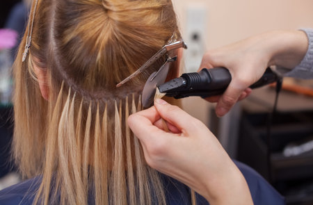 The hairdresser does hair extensions to a young girl, a blonde in a beauty salon. Professional hair...