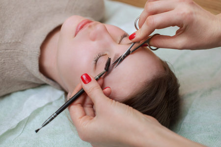 The make-up artist combs and shear her brows with scissors to a beautiful young girl in a beauty...