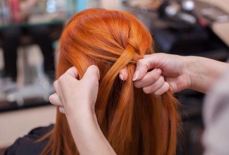 Beautiful red-haired girl, hairdresser weave a French braid close-up, in a beauty salon. Professional hair care and creating hairstyles.の写真素材