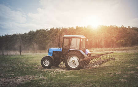 The tractor goes and pulls a plow plowing a field before landing of crops. In the early spring morning because of the wood the bright sun ascends.の写真素材
