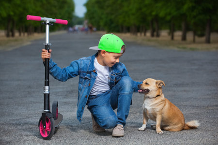 A small boy with a scooter sits next to a dog and strokes her in the park for a walk.の写真素材