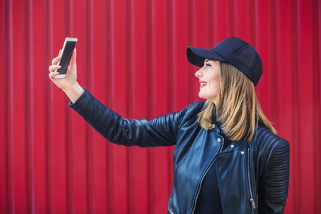Attractive blond girl in leather black jacket and cap, surfing the Internet on a mobile phone, on a...
