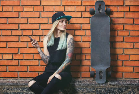 Beautiful, sexy hipster girl in tattoos against a red brick wall with a long board.の写真素材