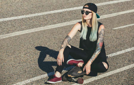 A beautiful, sexy hipster blonde with blue hair in tattoo sits on a longboard  with glasses and a cap on the asphalt on the streetの写真素材