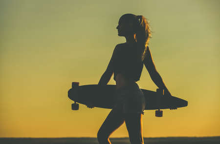 Silhouette of a beautiful, sexy hipster girl in tattoos standing with a longboard against the sunset at the sea.の写真素材