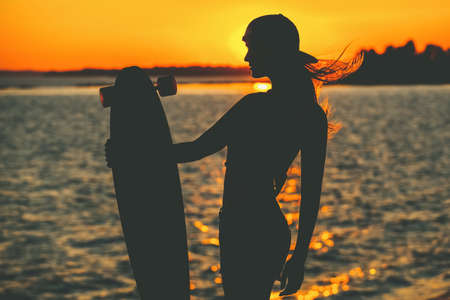 Silhouette of a beautiful, sexy hipster girl in tattoos standing with a longboard against the sunset at the sea.の写真素材
