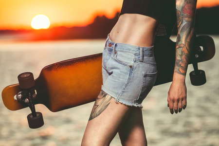 A beautiful, sexy hipster girl in tattoos stands with a longboard against the sunset at the sea.の写真素材