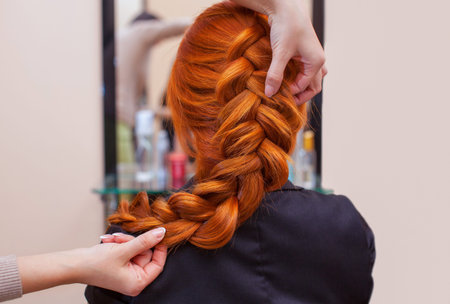 Beautiful, red-haired girl with long hair, hairdresser weaves a French braid, in a beauty salon....