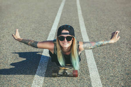 A beautiful, sexy blond hipster with blue hair in a tattoo is riding a longboard on an asphalt road.の写真素材