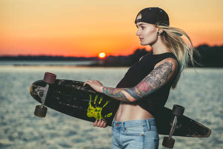 A beautiful, sexy hipster girl in tattoos stands with a longboard against the sunset at the sea.の写真素材