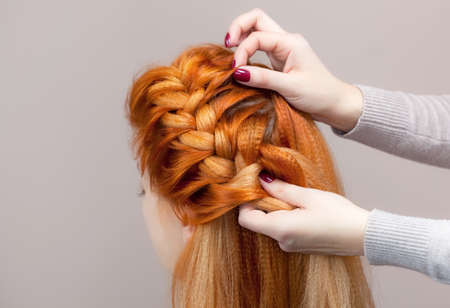 Beautiful, with long, red-haired hairy girl, hairdresser weaves a  braid, close-up in a beauty salon. Professional hair care and creating hairstyles.の写真素材