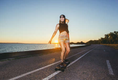 A beautiful sexy blond hipster with blue hair in a tattoo is riding a longboard on an asphalt road.の写真素材