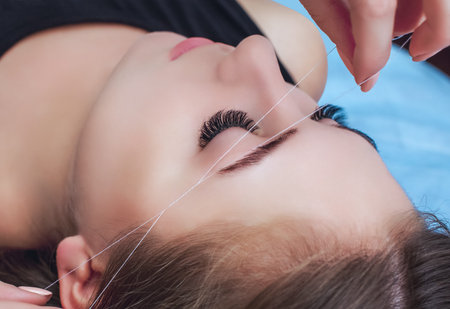 A make-up artist plucks an eyebrow with a thread from a beautiful brunette girl in a beauty salon.の写真素材