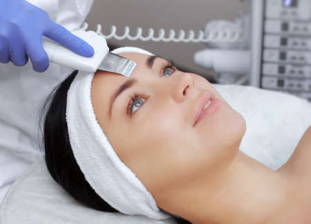 The doctor-cosmetologist makes the apparatus a procedure of ultrasound cleaning of the facial skin of a beautiful, young woman in a beauty salon. Cosmetology and professional skin care.の写真素材