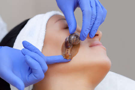 Cosmetological procedure. Beautiful young woman with a snail ahatin on her face in a beauty salon.の写真素材