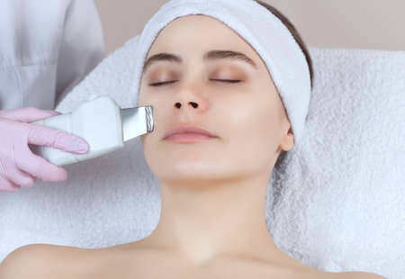 The doctor-cosmetologist makes the apparatus a procedure of ultrasound cleaning of the facial skin of a beautiful, young woman in a beauty salon. Cosmetology and professional skin care.の写真素材