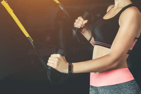 A slender girl holds a strap in her hand for the suspension training in the gym. TRX exercises. Sports concept and healthy lifestyle.の写真素材