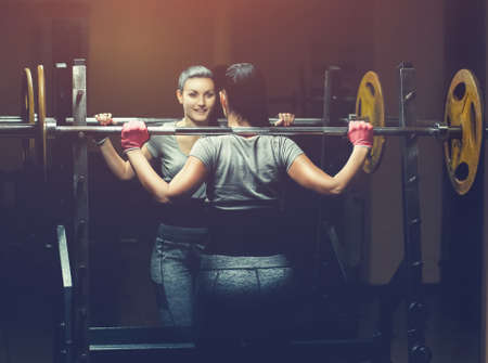 Slim girl bodybuilder lifting heavy barbell standing in front of the mirror while training in the gym. Sports concept, fat burning and a healthy lifestyle.の写真素材