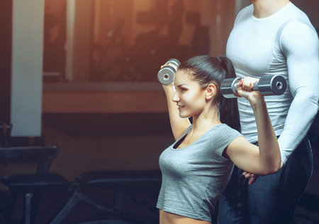 The coach helps a young athlete perform an exercise lifting dumbbells with a sports hall. Sports...