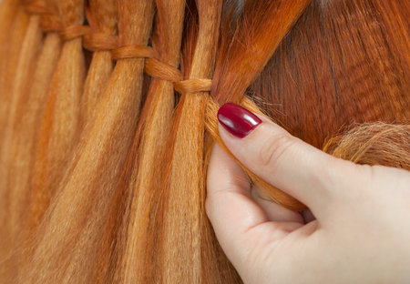 Beautiful girl with red hair, hairdresser weaves a  braid close-up, in a beauty salon. Professional...