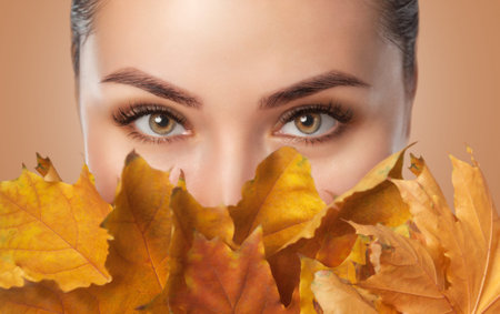 Beautiful woman with long eyelashes and with beautiful smokey eyes makeup holds yellow leaves near the face. Eyes close up. Looking at the cameraの写真素材