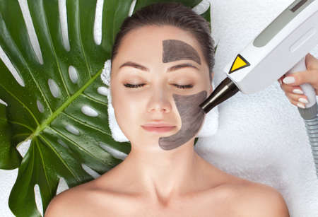 Cosmetologist does Carbon face peeling procedure of a beautiful, young woman in a beauty salon. In the background lies a leaf of a monstera plant. Hardware cosmetology treatment and Spa.の写真素材