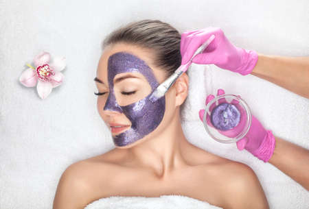 Beautician makes a face mask with purple particles on the face of a woman to rejuvenate the skin. Cosmetology treatment of problem skin on the face and body.の写真素材