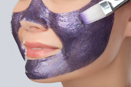 Beautician makes a face mask with purple particles on the face of a woman to rejuvenate the skin. Cosmetology treatment of problem skin on the face and body.の写真素材