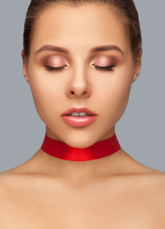 Beautiful woman with clean skin and pink makeup, with closed eyes with a red ribbon on her neck.  Makeup and cosmetology concept.の写真素材
