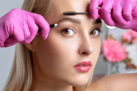 Eyelash artist plucks eyebrows with tweezers. Beautiful blonde woman having Permanent Make-up Tattoo on her Eyebrows. Professional makeup and cosmetology skin care.の写真素材