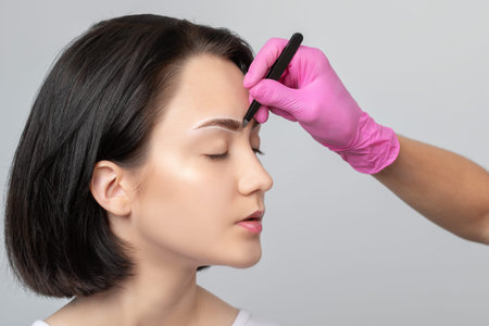 Brunette teenager girl with dark hair having Permanent Make-up Tattoo on her Eyebrows. Eyelash artist plucks eyebrows with tweezers. Professional makeup and cosmetology skin care.の写真素材