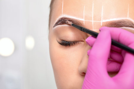 teenager girl having permanent makeup tattoo on her eyebrows. Make-up artist makes markings with...