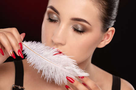 Portrait of a beautiful girl with dark hair, with beautiful makeup and healthy clean skin. She is holding a white peacock feather near her face. Makeup and cosmetology concept.の写真素材
