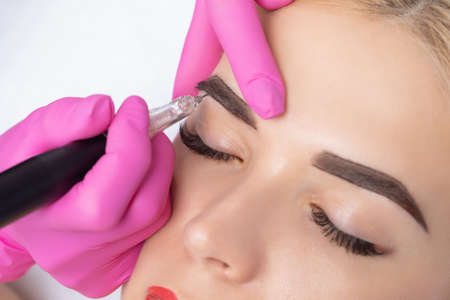 Permanent make-up for eyebrows of beautiful blonde woman in beauty salon. Closeup beautician doing eyebrows tattooing.の写真素材