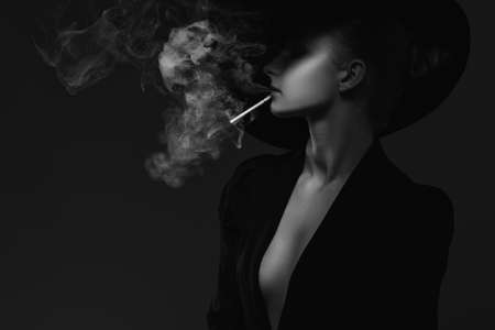 A mysterious woman in a black suit and hat smokes a cigarette. black and white photo.の写真素材