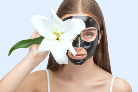 Carbon peeling procedure to beautiful blue-eyed girl in a beauty salon. Hardware cosmetology treatment.A girl with a black mask on her face holds a white lily.の写真素材