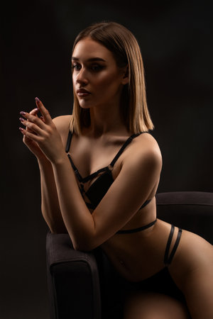 A slender woman in a black swimsuit sits on a chair on a dark background.の写真素材