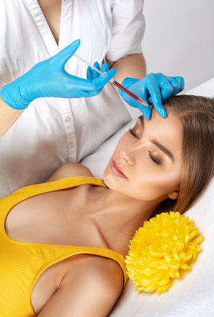 Cosmetologist makes rejuvenating anti wrinkle injections on the face of a beautiful woman in a yellow blouse. Female aesthetic cosmetology in a beauty salon.の写真素材