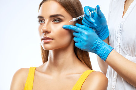 Cosmetologist makes rejuvenating anti wrinkle injections on the face of a beautiful woman in a yellow blouse. Female aesthetic cosmetology in a beauty salon.の写真素材