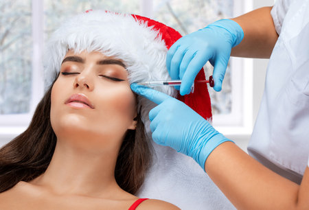 The cosmetologist makes anti-aging anti-wrinkle injections on the face of a beautiful woman in a red santa hat. Women's aesthetic cosmetology in a beauty salon. New year concept.の写真素材