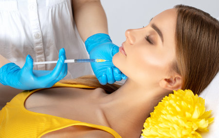 Cosmetologist makes rejuvenating anti wrinkle injections on the face of a beautiful woman in a yellow blouse. Female aesthetic cosmetology in a beauty salon.の写真素材