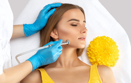 A cosmetologist doctor performs a microdermabrasion procedure on the face against acne and blackheads in a beautiful young girl in yellow. Women's cosmetology in a beauty salon.の写真素材