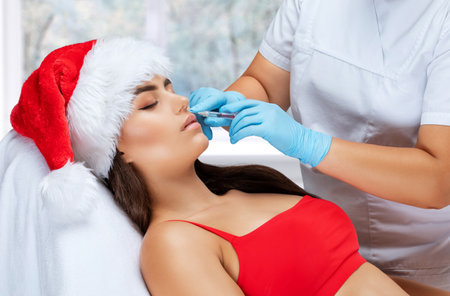 The cosmetologist makes anti-aging anti-wrinkle injections on the face of a beautiful woman in a red santa hat. Women's aesthetic cosmetology in a beauty salon. New year concept.の写真素材