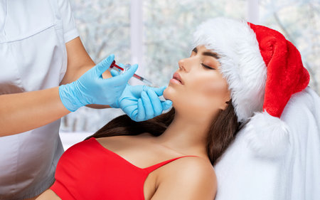 The cosmetologist makes anti-aging anti-wrinkle injections on the face of a beautiful woman in a red santa hat. Women's aesthetic cosmetology in a beauty salon. New year concept.の写真素材