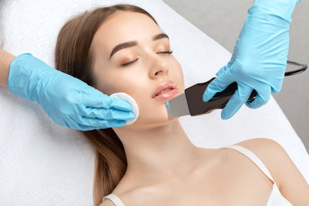 The doctor-cosmetologist makes the ultrasound cleaning procedure of the facial skin of a beautiful, young woman in a beauty salon. Cosmetology and professional skin care.の写真素材
