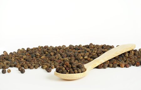 Black pepper and wooden spoon on white background. Spoon in front of heap of grains of pepper....