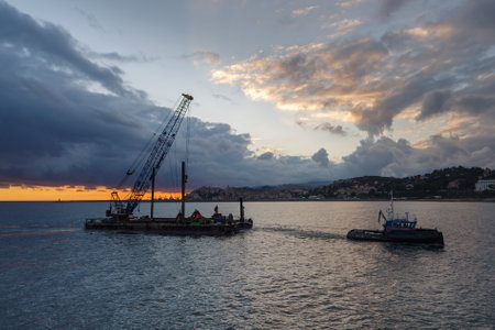 Tugboat pulls a construction crane barge beside the coastline during land reclamation work off the coast of Liguria, Italyの写真素材