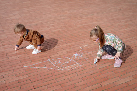 Girl with her younger brother doodling with chalk on pavement, overhead view